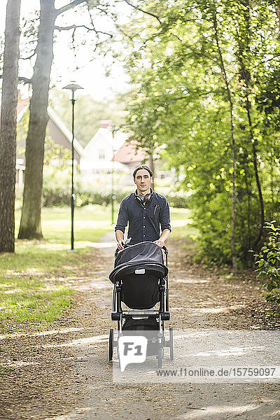 Portrait of man walking while pushing baby carriage on footpath at public park