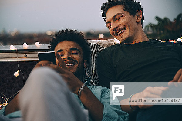 Happy male friends looking at smart phone relaxing on terrace during rooftop party in city