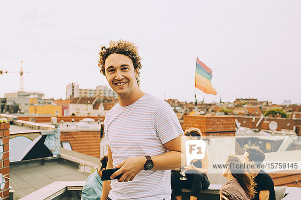 Porträt eines lächelnden jungen Mannes mit Mobiltelefon  während sich Freunde während einer Party auf der Terrasse vergnügen