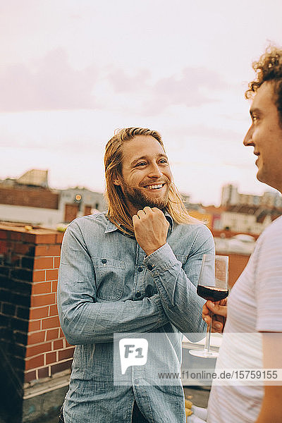 Smiling male friends talking while standing on terrace at rooftop party