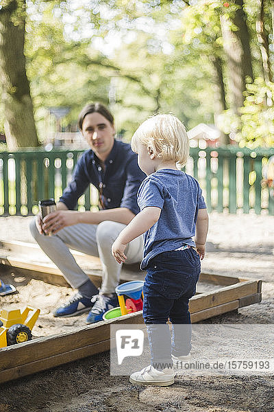 Mann sitzt  während er den auf dem Spielplatz stehenden Sohn betrachtet