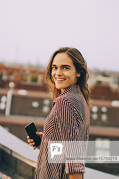 Portrait of happy young woman holding mobile phone standing on terrace in city
