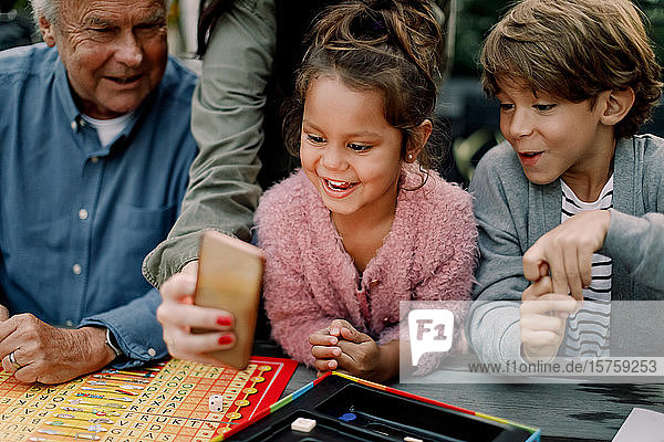 Cropped hand of woman showing mobile phone to children and senior man sitting at table