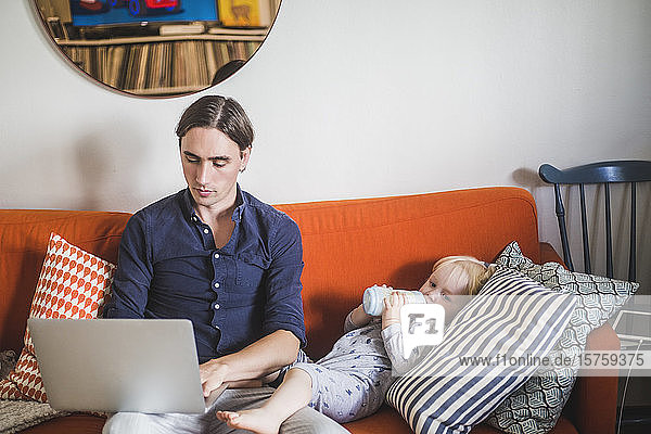 Stay at home father using laptop while sitting by toddler son drinking milk from bottle on sofa in apartment