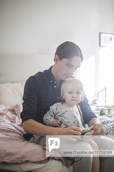 Father assisting baby boy wearing clothes while sitting on bed at home