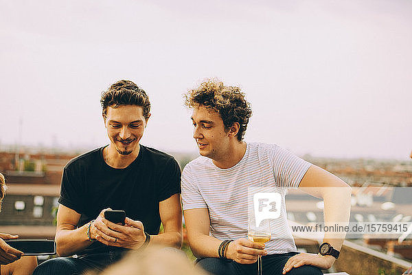 Mann zeigt einem Freund sein Handy und trinkt während der Party auf der Terrasse