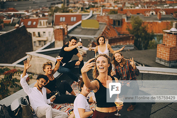 Frau trinkt  während sie sich mit Freunden am Smartphone auf der Terrasse bei einer Party einen Drink genehmigt