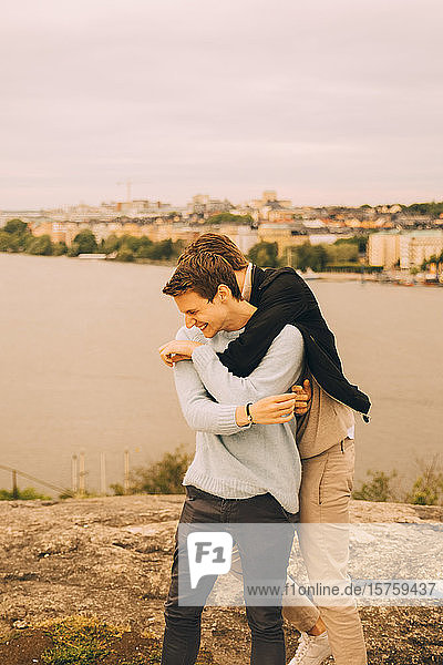 Cheerful male friends embracing while standing on rock formation against lake