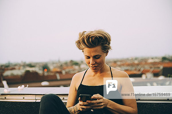 Lächelnde Frau mit Kurzhaar-SMS auf Smartphone  während sie auf der Terrasse gegen den Himmel sitzt