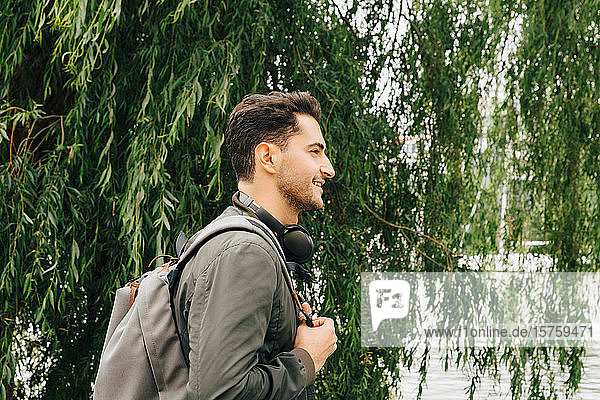 Seitenansicht eines jungen Mannes mit Rucksack  der in der Stadt an einem Baum steht