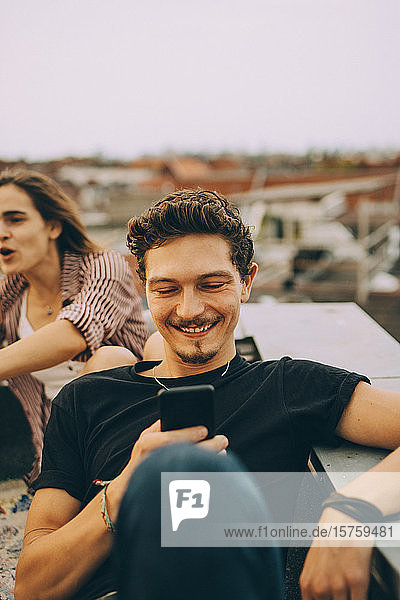 Junger Mann benutzt Smartphone  während ein Freund auf einer Dachparty feiert