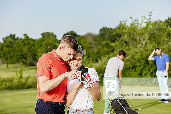 Couple using smartphone on golf course  friends playing golf in background