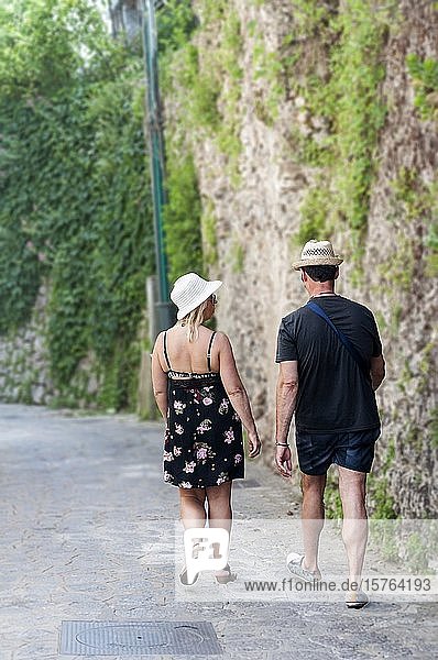 A man and a woman dressed in casual summer clothes walk down a typical street near Ravello on the Amalfi Coast. Stone walls covered in green vines are a common site all over the Amalfi Coast.