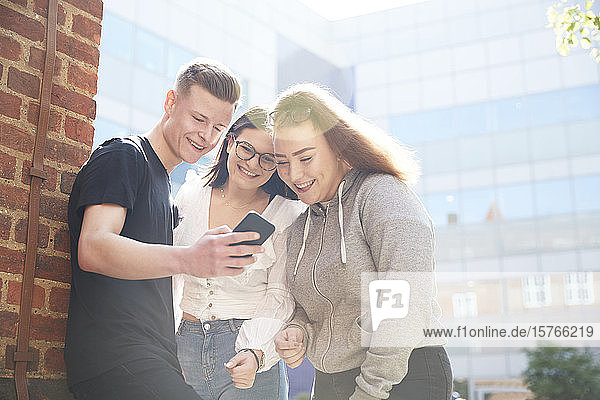 Freunde im Teenageralter benutzen ein Smartphone vor einem sonnigen Schulgebäude