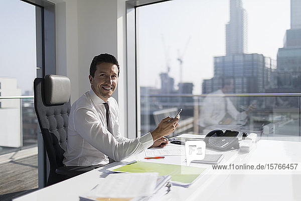Porträt eines selbstbewussten Geschäftsmannes  der ein Smartphone in einem Hochhausbüro benutzt