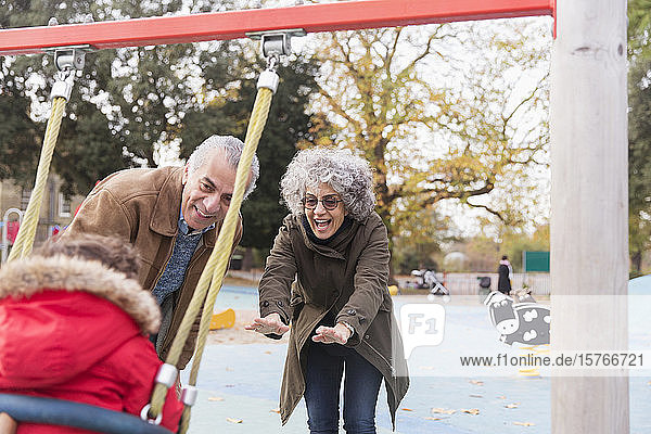 Verspielte Großeltern schieben ihren kleinen Enkel auf dem Spielplatz auf eine Schaukel