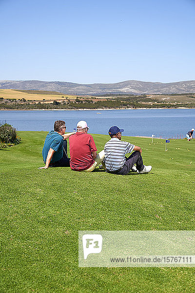 Männliche Golfer machen eine Pause und ruhen sich im Gras auf einem sonnigen Golfplatz aus
