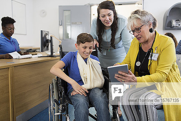 Ärztin mit digitalem Tablet im Gespräch mit einer Mutter und einem kleinen Patienten im Rollstuhl in der Kliniklobby