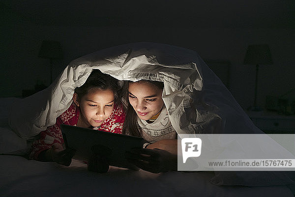 Schwestern schauen einen Film auf einem digitalen Tablet unter einer Decke im dunklen Schlafzimmer