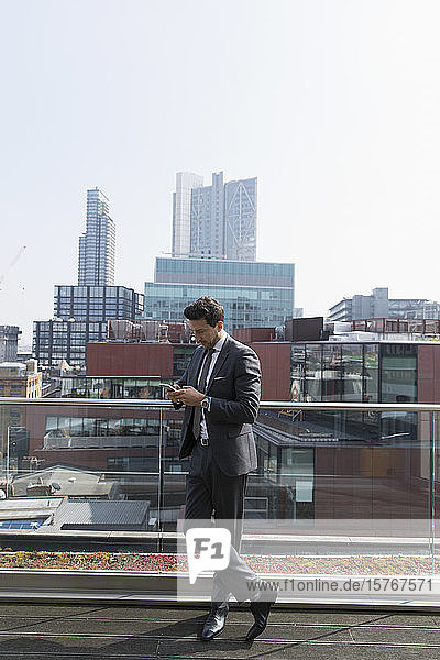 Geschäftsmann  der auf einem sonnigen  städtischen Hochhausbalkon ein Smartphone benutzt