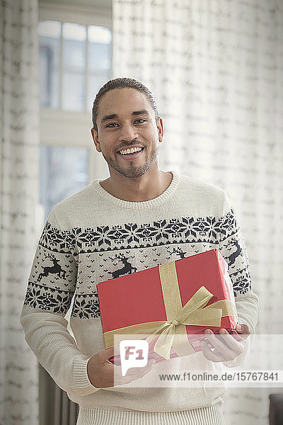 Porträt lächelnder junger Mann im Weihnachtspulli mit Geschenk