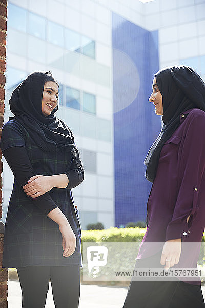 Junge Freundinnen in Hijabs unterhalten sich vor einem sonnigen Gebäude