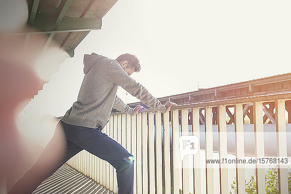 Junger männlicher Läufer  der sein Bein an einem sonnigen städtischen Geländer ausstreckt