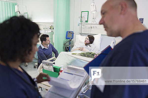 Ärzte mit Krankenblatt machen Visite mit einem Patienten  der im Krankenhausbett im Hintergrund ruht