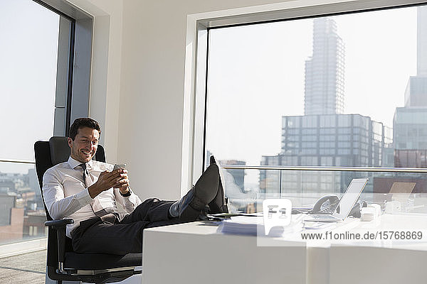 Lächelnder Geschäftsmann  der ein Smartphone benutzt und die Füße auf den Schreibtisch legt  in einem modernen  sonnigen  städtischen Büro