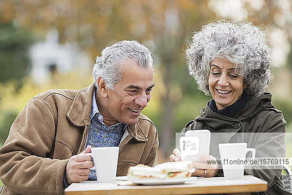 Älteres Paar mit Smartphone  das im Park zu Mittag isst und Kaffee trinkt