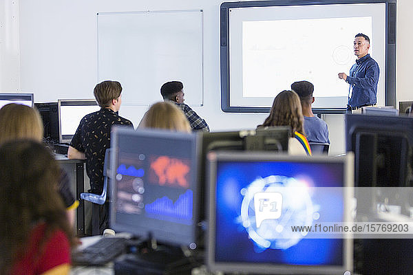 Schüler der Mittelstufe an Computern  die den Lehrer auf der Projektionsfläche im Klassenzimmer beobachten