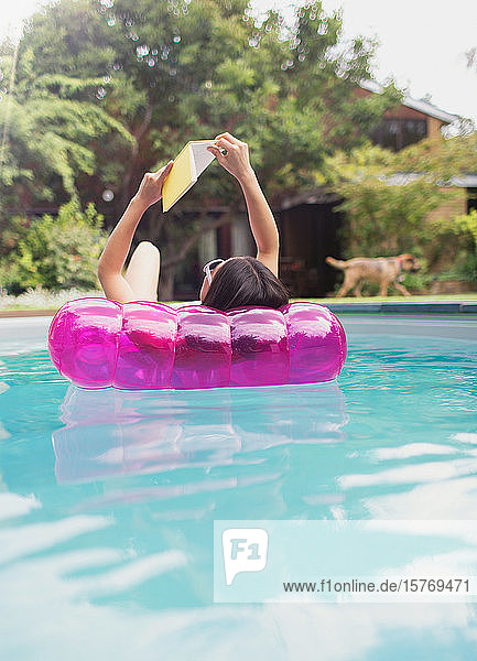 Woman relaxing reading book on inflatable raft in sunny summer swimming pool