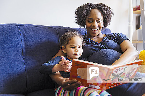 Schwangere Mutter liest ihrer Tochter auf dem Sofa ein Buch vor