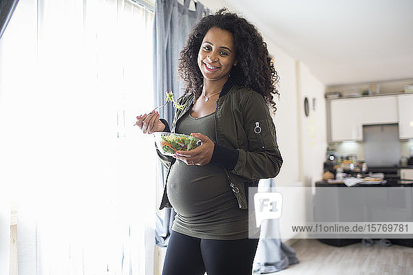 Porträt glückliche junge schwangere Frau isst Salat am Fenster