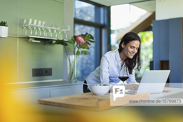 Smiling woman using laptop and drinking red wine in kitchen