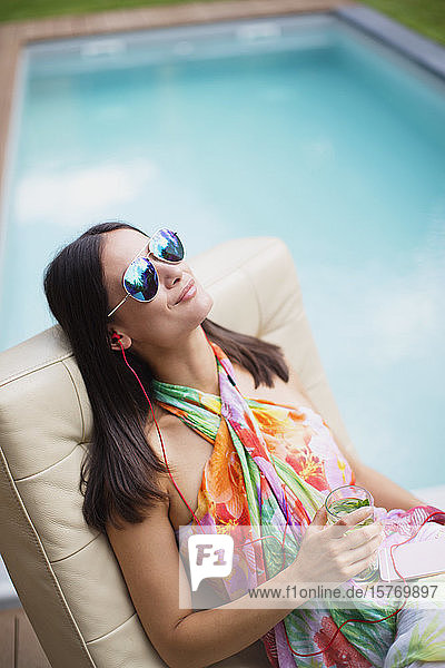 Gelassene Frau mit Kopfhörern und Cocktail  die sich am Pool entspannt und Musik hört