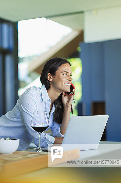 Lächelnde Frau  die am Laptop mit einem Smartphone spricht und Rotwein trinkt