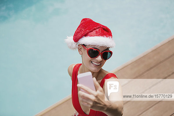 Verspielte Frau mit Weihnachtsmannmütze und herzförmiger Sonnenbrille macht Selfie mit Fotohandy am Pool