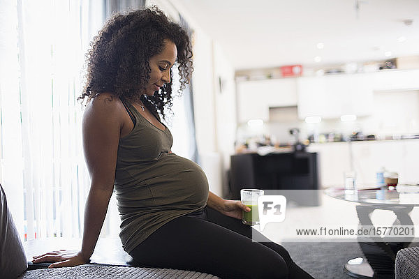 Junge schwangere Frau trinkt grünen Smoothie im Wohnzimmer