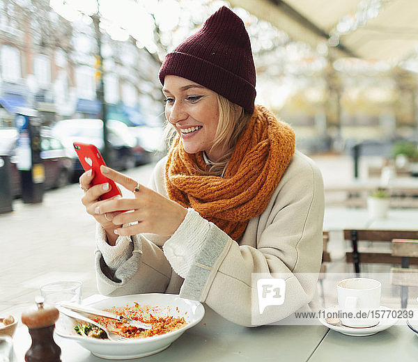 Lächelnde junge Frau mit Smartphone beim Mittagessen im Herbst in einem Straßencafé