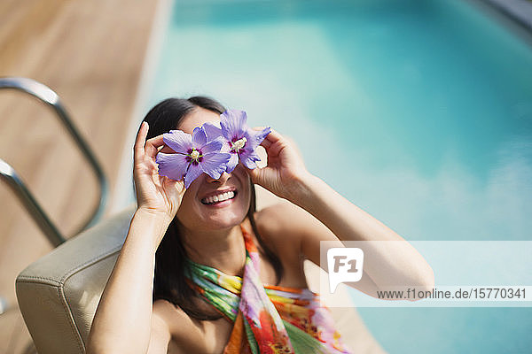 Portrait playful woman holding purple hibiscus over eyes at sunny summer poolside