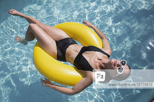 Sinnliche Frau im schwarzen Bikini schwimmt auf aufblasbarem Ring im sonnigen Sommerschwimmbad