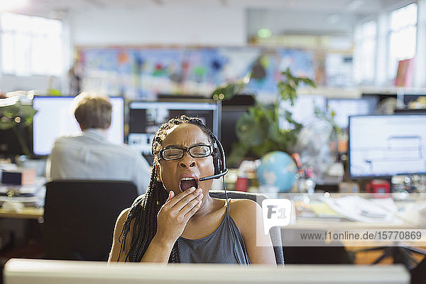 Müde Geschäftsfrau mit Headset gähnt am Computer im Großraumbüro