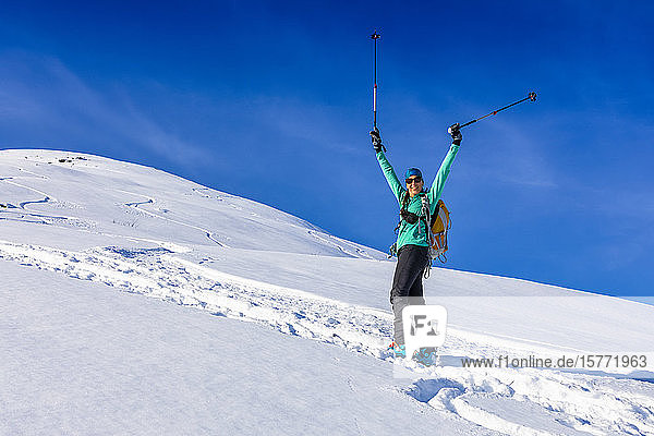 Frau beim Skilanglauf  die einen schönen Tag mit ihren Stöcken in der Luft feiert und mit AT-Skiern und Fellen in Hatcher's Pass  Alaska  Talkeetna Mountains  Alaska  Vereinigte Staaten von Amerika  den Berg hinaufklettert