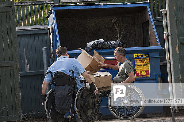 Männer in Rollstühlen  die Kartons in eine Mülltonne werfen