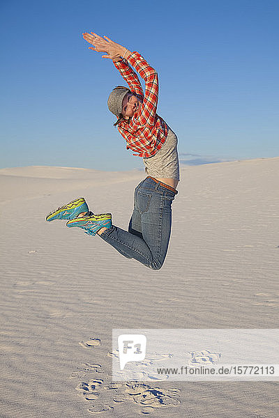 A young woman leaps in the air on the white sand with blue sky White Sands National Monument; Alamogordo New Mexico United States of America