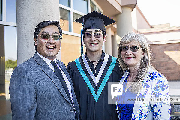A young man wearing a cap and gown stands with his parents for a portrait on graduation day; Surrey  British Columbia  Canada