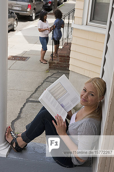 A young woman reading from a textbook on her front porch and looking up at the camera  with a mother and daughter in the background in front of their house