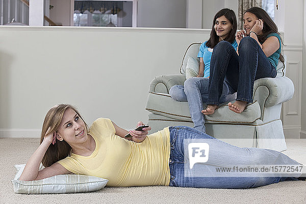 Drei Mädchen im Teenageralter sitzen zusammen zu Hause  eine sieht fern und wechselt den Kanal mit der Fernbedienung  die anderen sitzen auf einem Stuhl und hören Musik