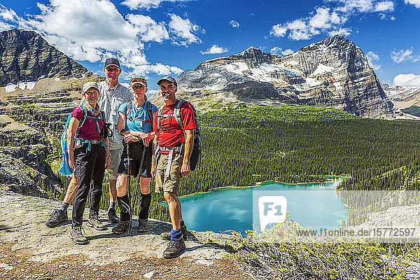 Two sets of couples hikers standing on rocky ridge with blue alpine lake and mountains in the distance with blue sky and clouds  Yoho National Park; Field  British Columbia  Canada